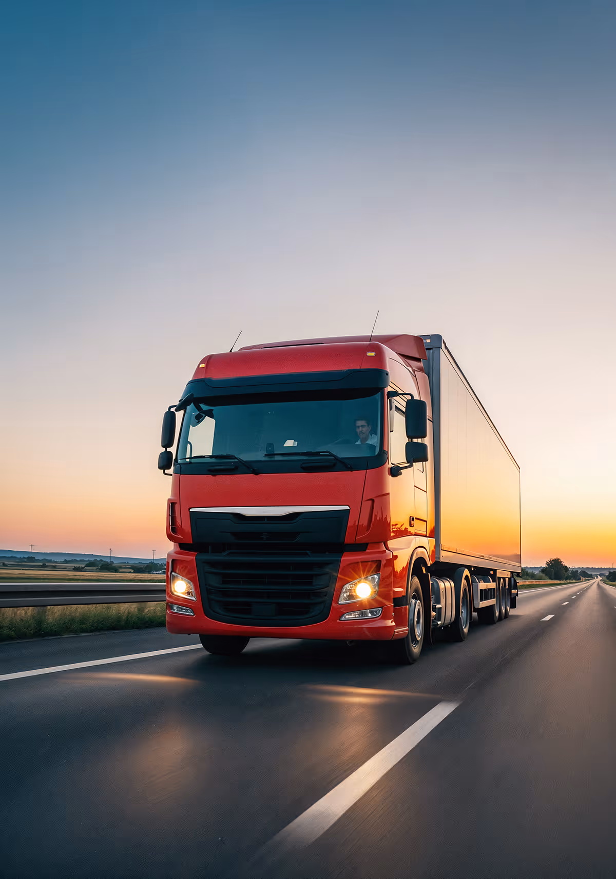 Red semi-truck driving on a highway at sunset with headlights on.